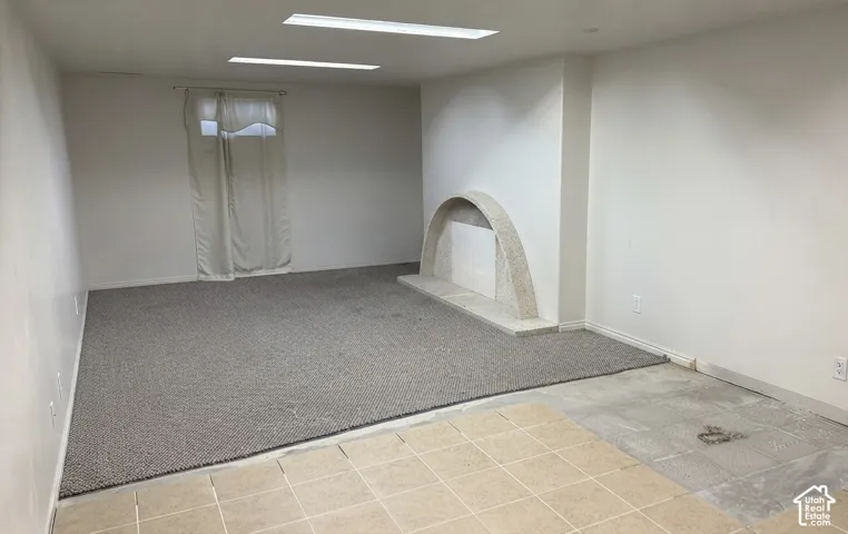 Spacious room featuring a light-colored carpet, a window with curtains, and a decorative fireplace with an arched opening