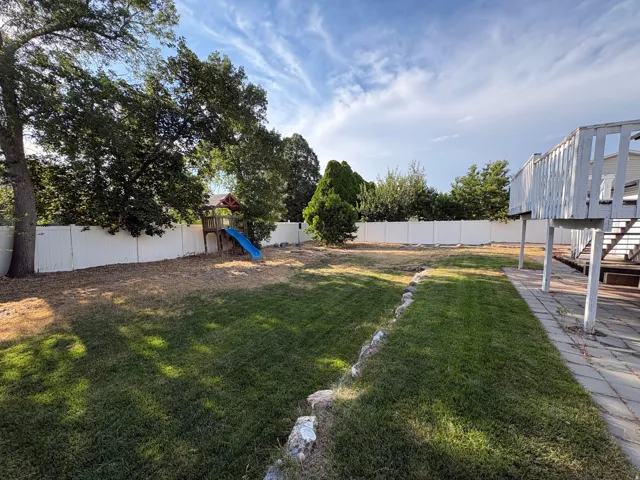 Expansive backyard featuring a white privacy fence, mature trees, a wooden deck with railing, and a paver stone patio