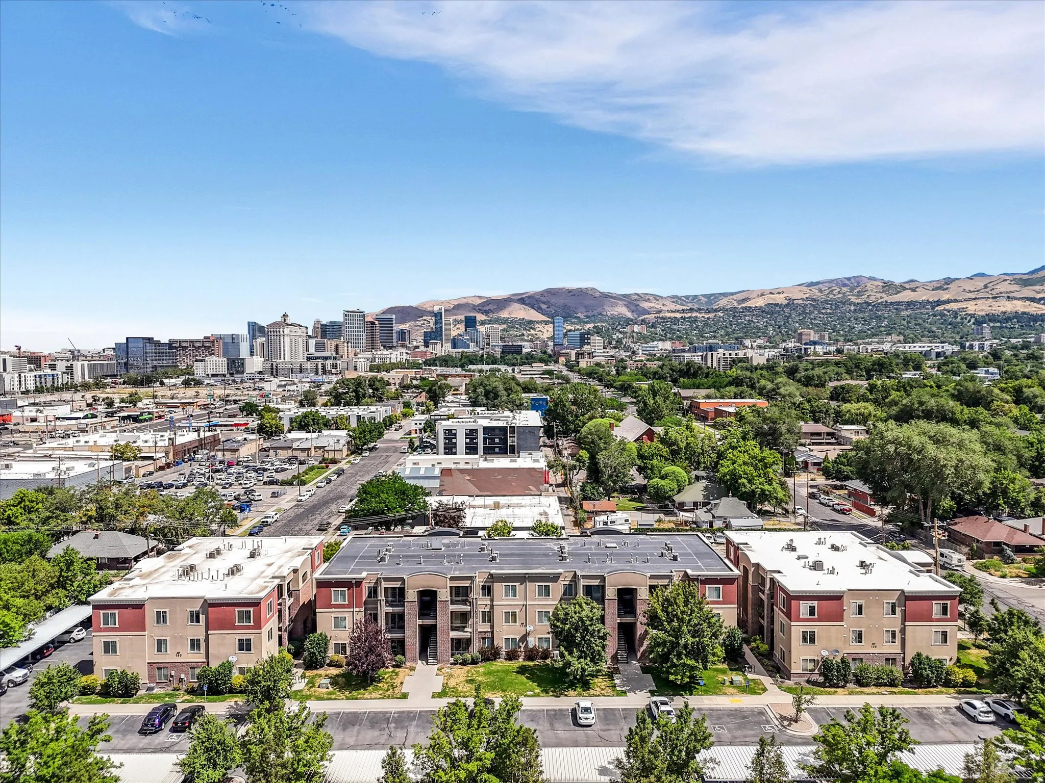 Expansive aerial capture showcasing multiple residential buildings, lush green trees, and distant city skyline against a mountainous backdrop