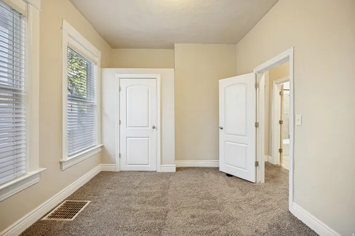 Carpeted room featuring two windows with horizontal blinds, a white paneled door, and a second open doorway revealing a partial bathroom beyond