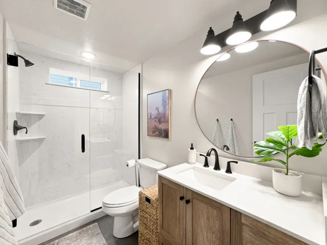 Glass-enclosed shower with a white tile surround and recessed window, wood-finish vanity with a white countertop and undermount sink, round mirror, and a three-light vanity fixture