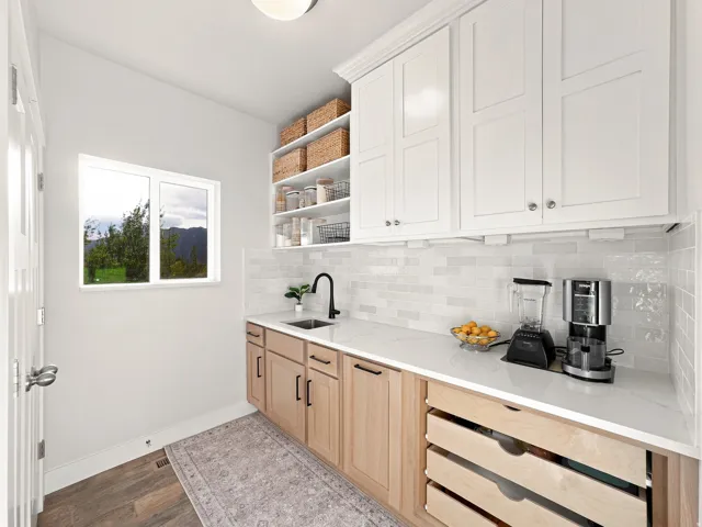White upper cabinetry, light wood-finish lower cabinetry, white subway tile backsplash, white countertop, and a single basin sink with a matte black faucet