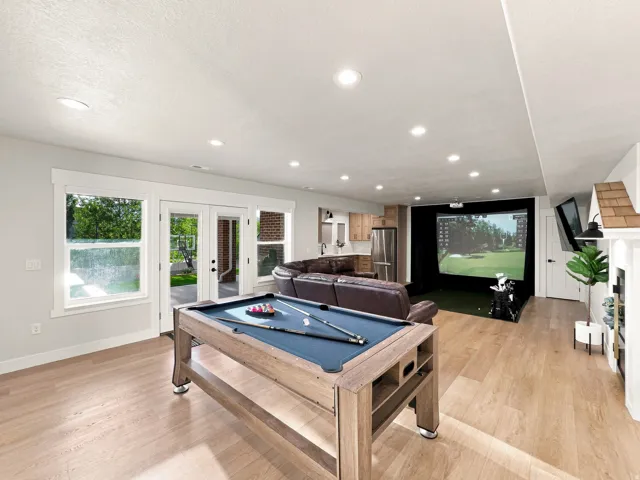 Recessed lighting throughout, wood-finish flooring, built-in wet bar with stainless steel refrigerator, white fireplace with decorative mantel, and a golf simulator screen