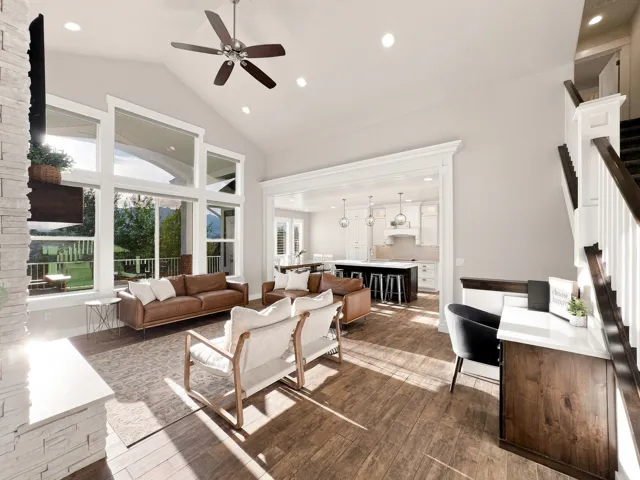 Vaulted ceiling with a ceiling fan, extensive window array, wood-finish flooring, stone-clad fireplace surround, and a built-in desk