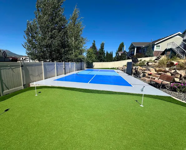 Backyard featuring a synthetic turf putting green with two flags, a multi-sport court with blue surfacing, and a rockery with integrated landscaping