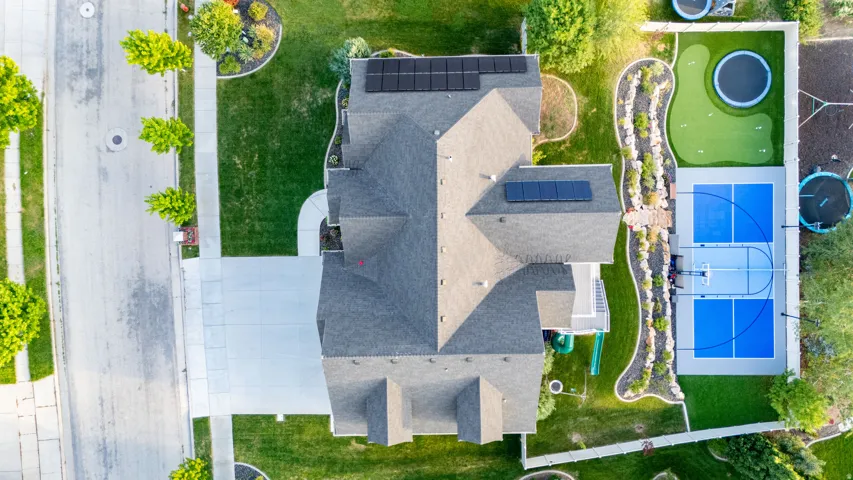Asphalt shingle roof with solar panels, concrete driveway, manicured lawn, and a multi-sport court with a putting green