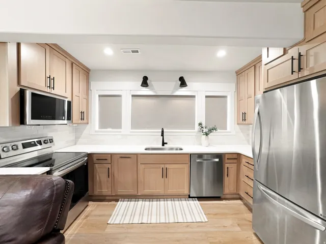 Wood-finish flooring, light wood cabinetry with black hardware, white countertops, subway tile backsplash, and stainless steel appliances