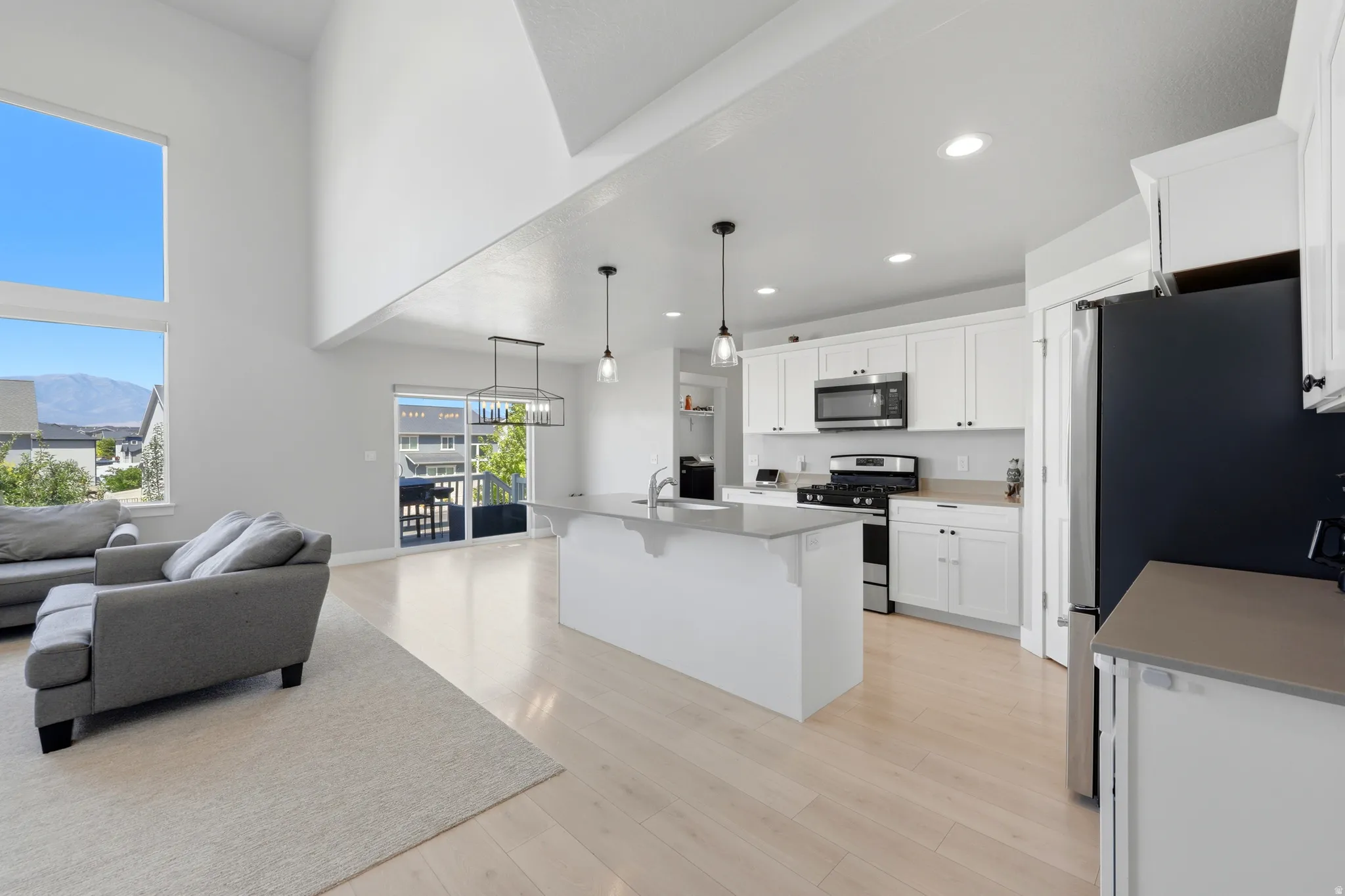 Open-concept living area featuring a two-story ceiling, large windows, light wood-finish flooring, and a contemporary kitchen with white cabinetry and an island