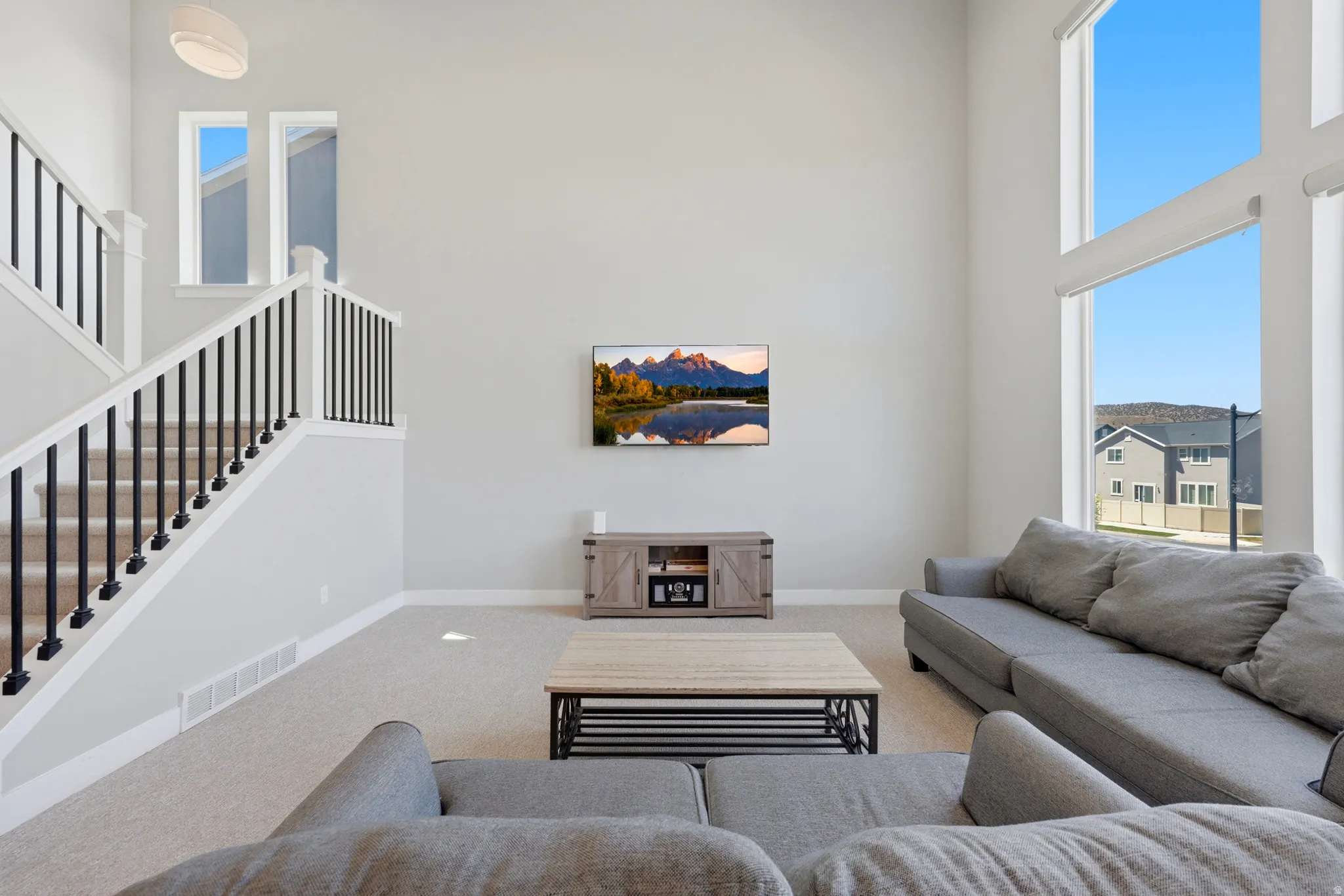 Two-story living area featuring a prominent staircase with black metal balusters and white risers, expansive two-story windows with roller shades, and light-colored carpeting