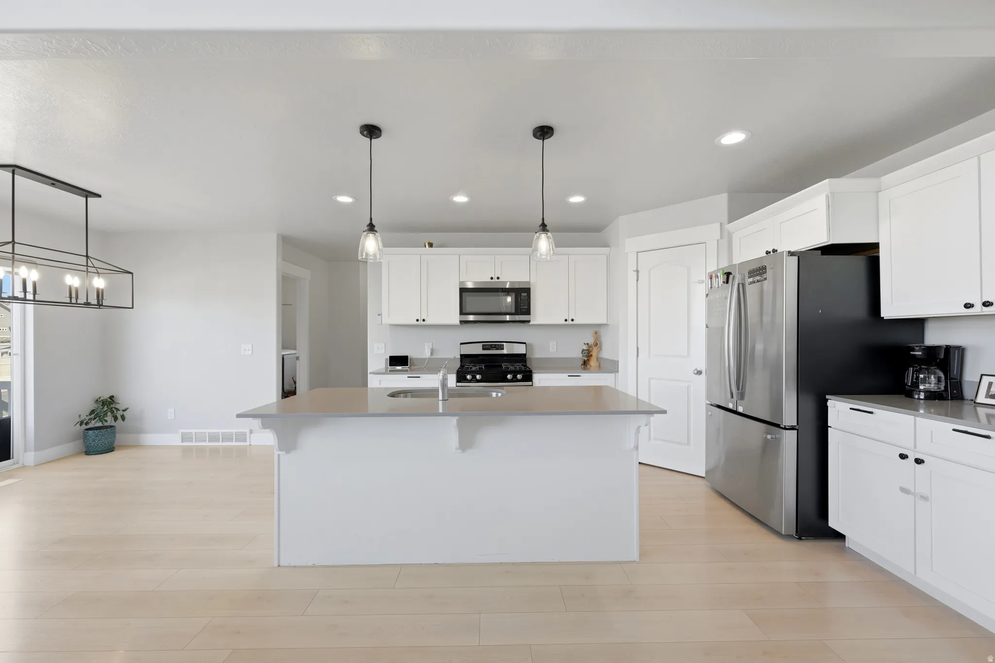 Open-concept kitchen featuring an oversized island with pendant lighting, light wood-finish flooring, white cabinetry with black hardware, recessed lighting, and stainless steel appliances