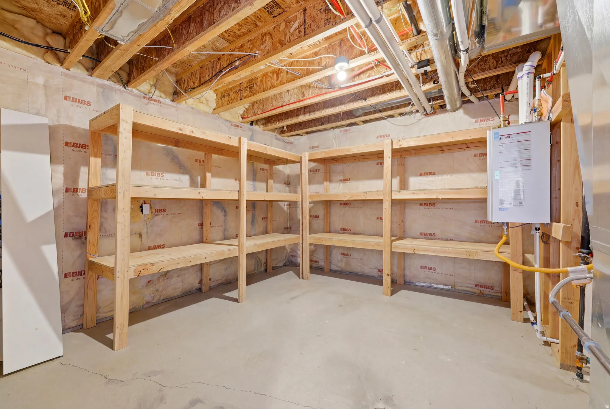 Unfinished basement area featuring exposed ceiling joists, concrete flooring, built-in wooden shelving, and a tankless water heater