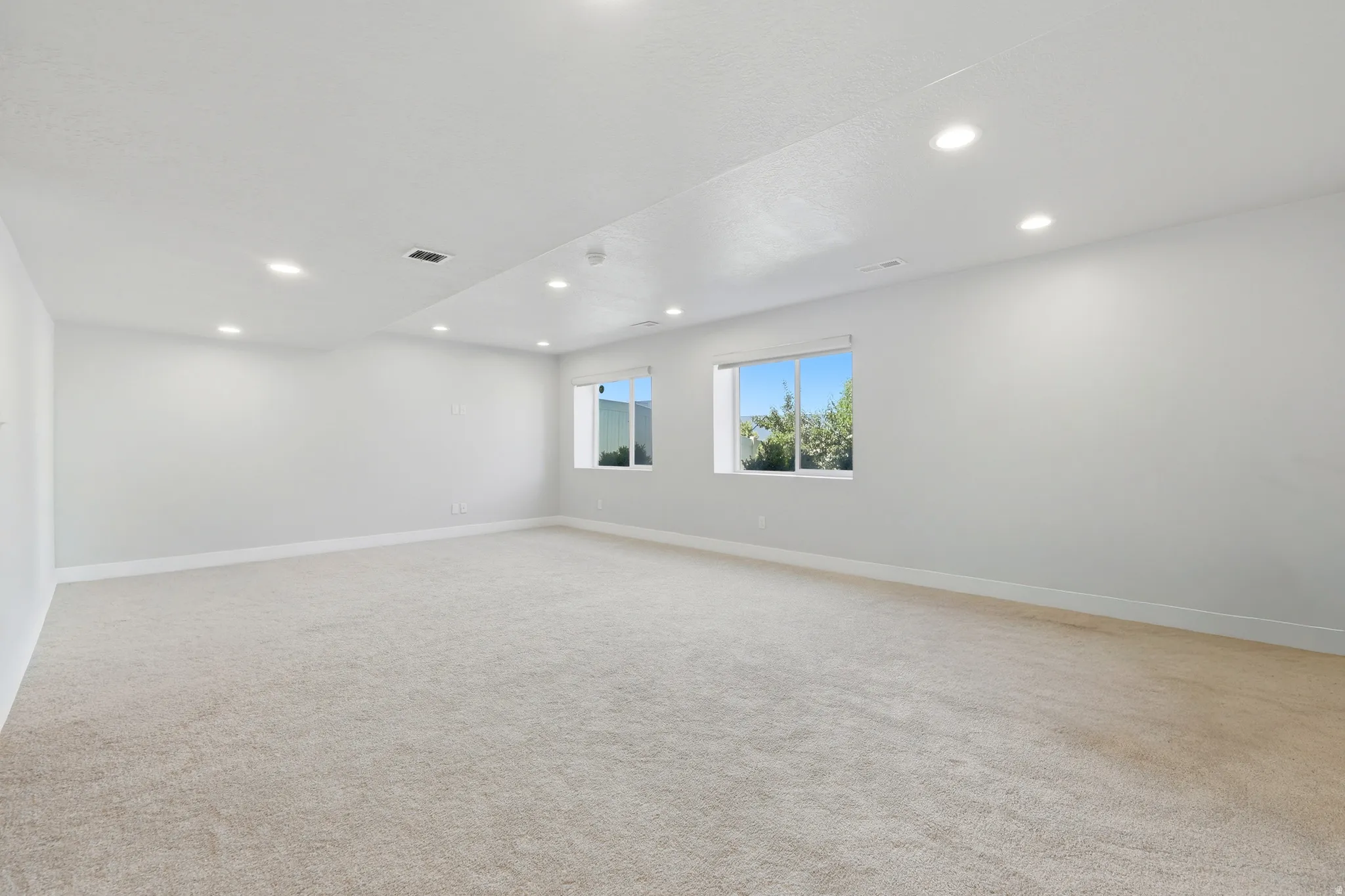 Spacious room with light-colored carpeting, recessed lighting, and multiple windows providing natural illumination