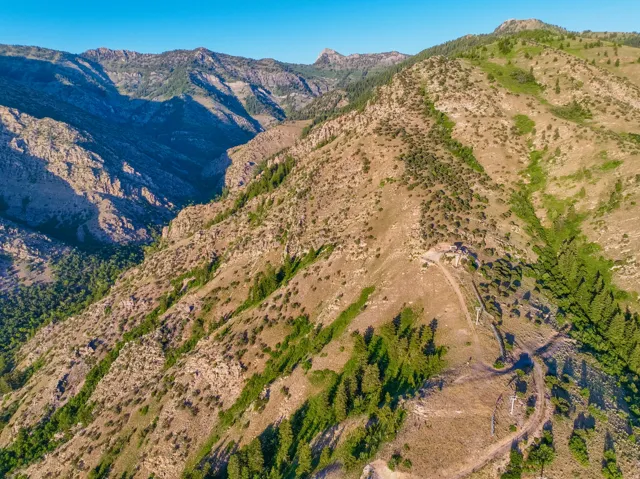 Expansive mountain landscape featuring rugged terrain, varied vegetation including evergreen trees, and a clear blue sky overhead