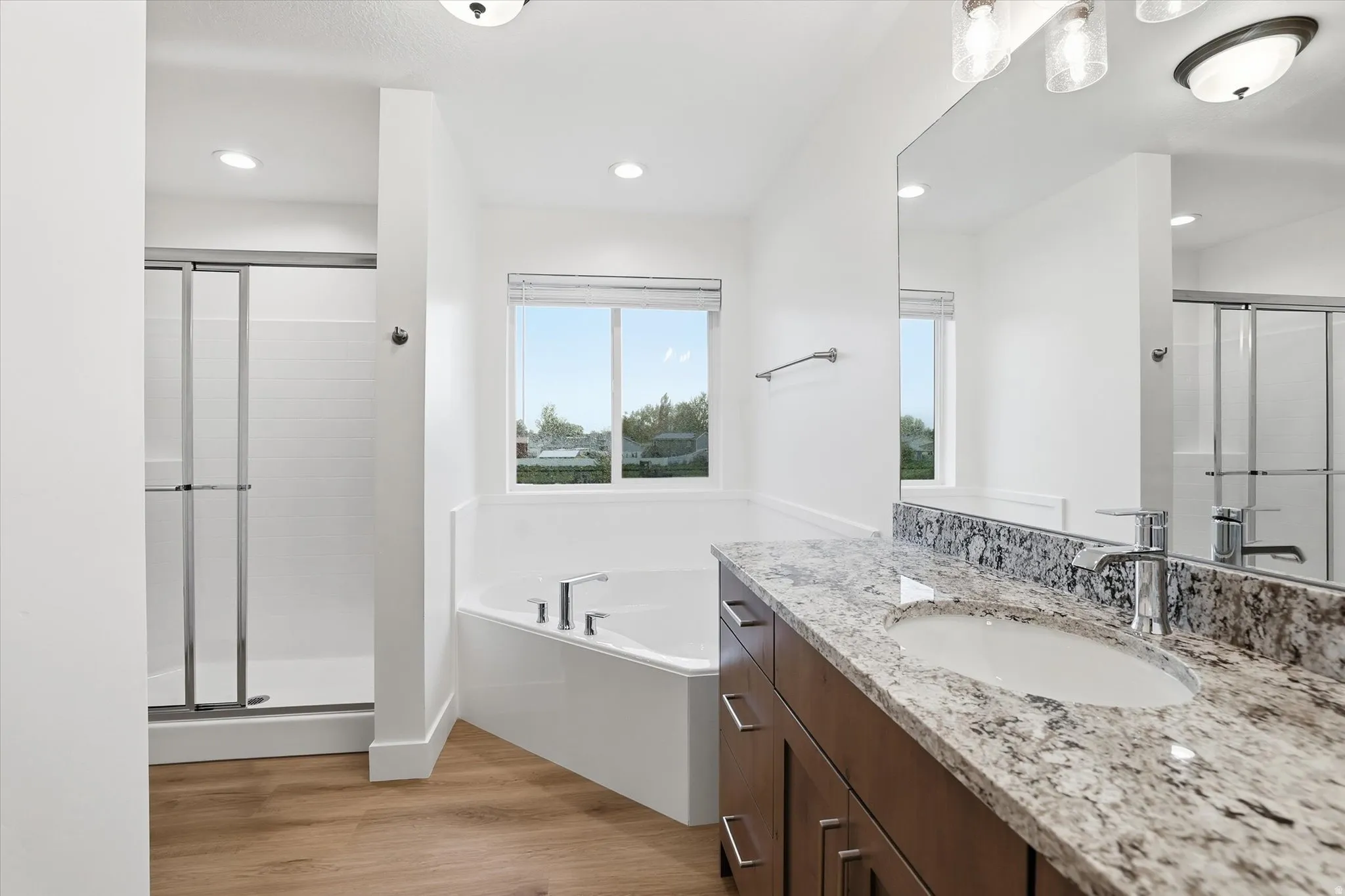 Bathroom featuring a double vanity with granite countertop, wood-finish flooring, a soaking tub, and a separate glass-enclosed shower with subway tile surround