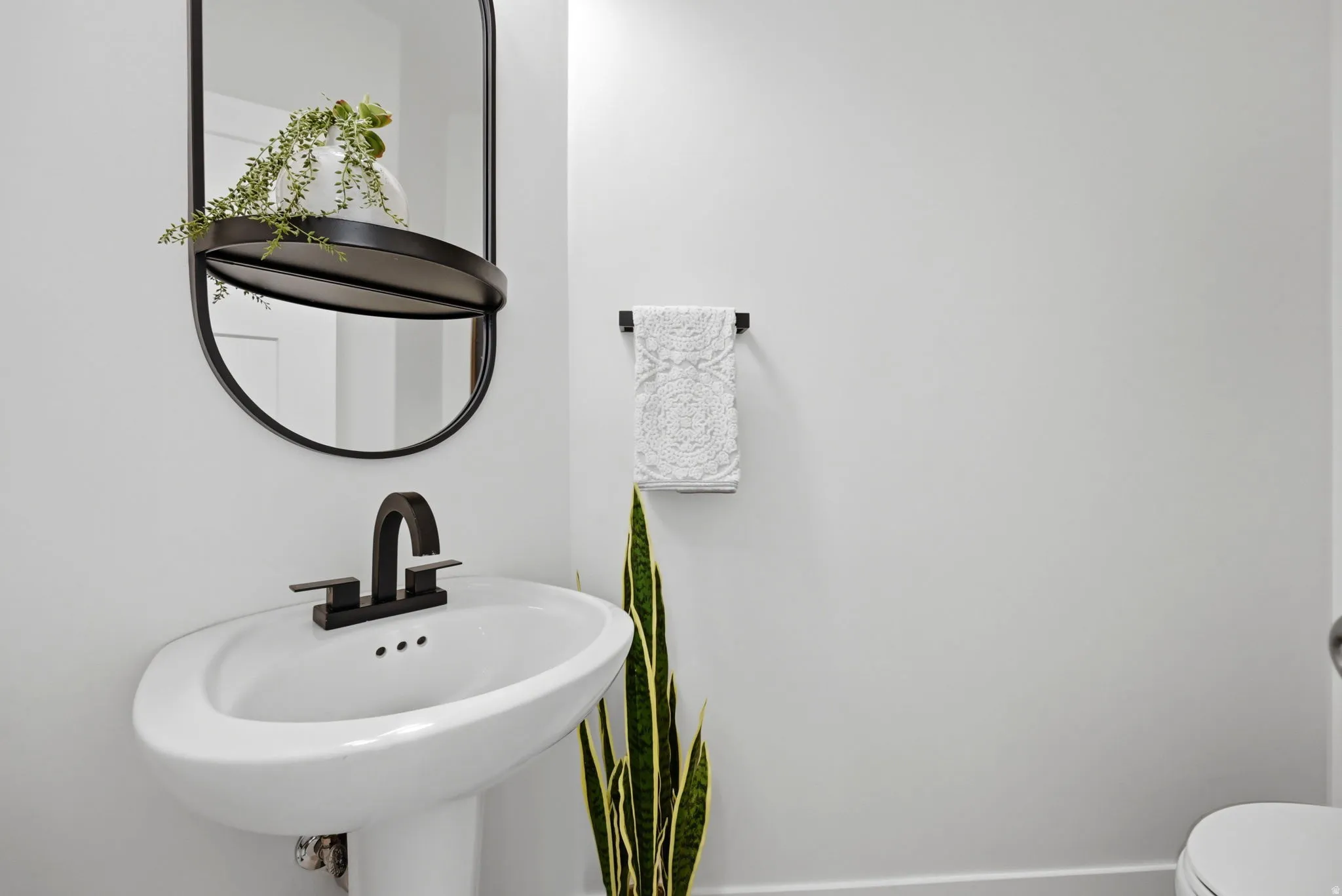 Pedestal sink with a matte black faucet and coordinating oval mirror featuring an integrated shelf