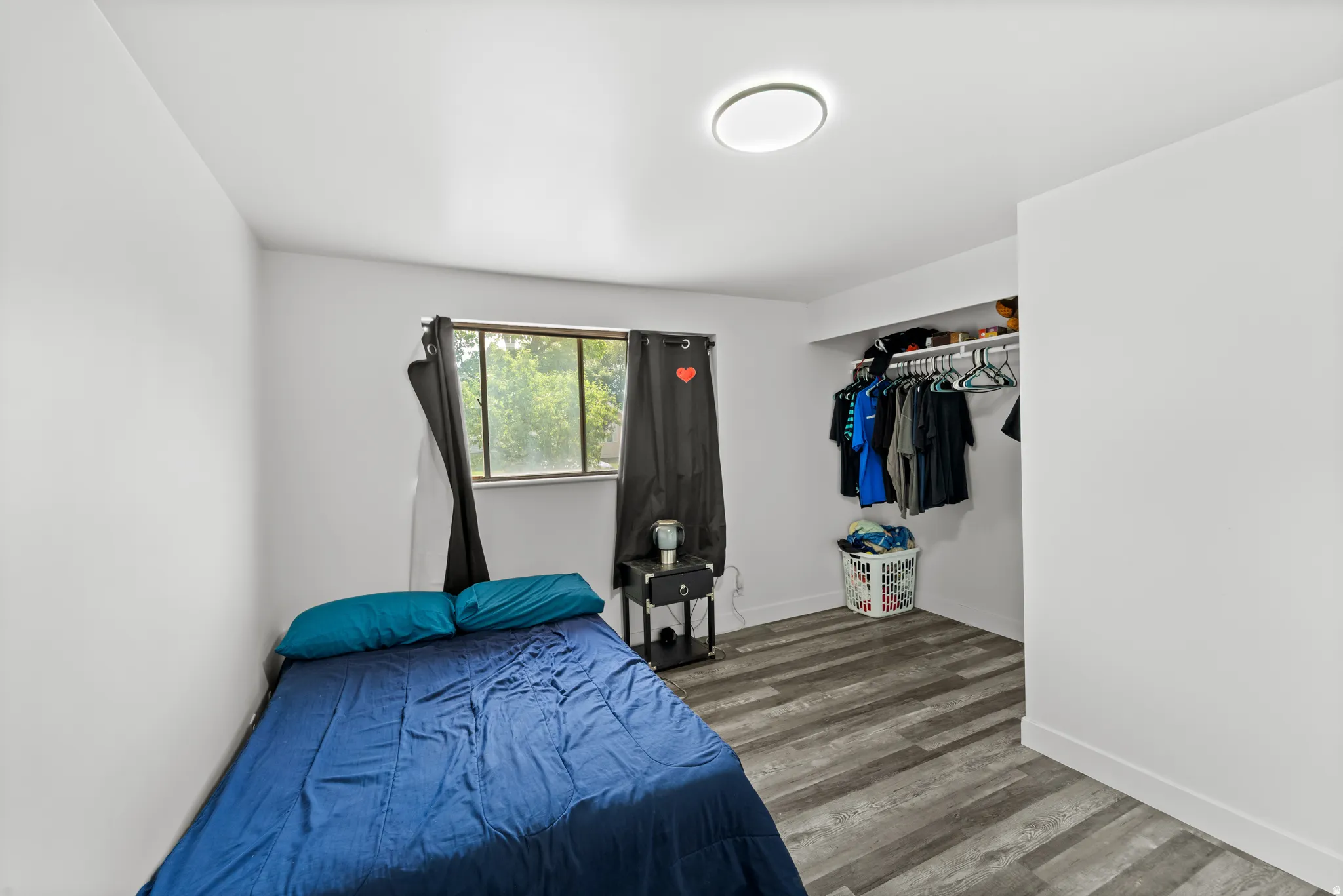 Bedroom featuring wood-finish flooring, a window with black curtains, an integrated closet with shelving and hanging rod, a ceiling-mounted light fixture, and white walls