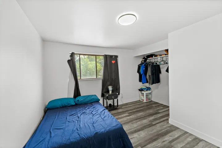 Bedroom featuring wood-finish flooring, a window with black curtains, an integrated closet with shelving and hanging rod, a ceiling-mounted light fixture, and white walls