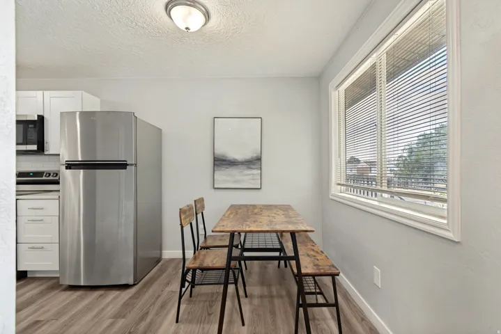 Kitchen and dining area featuring stainless steel appliances, wood-finish flooring, white cabinetry, a large window, and a ceiling-mounted light fixture