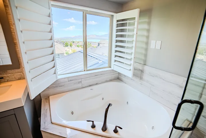 Soaking tub with bronze fixtures, surrounded by wood-look tile and a window featuring white plantation shutters