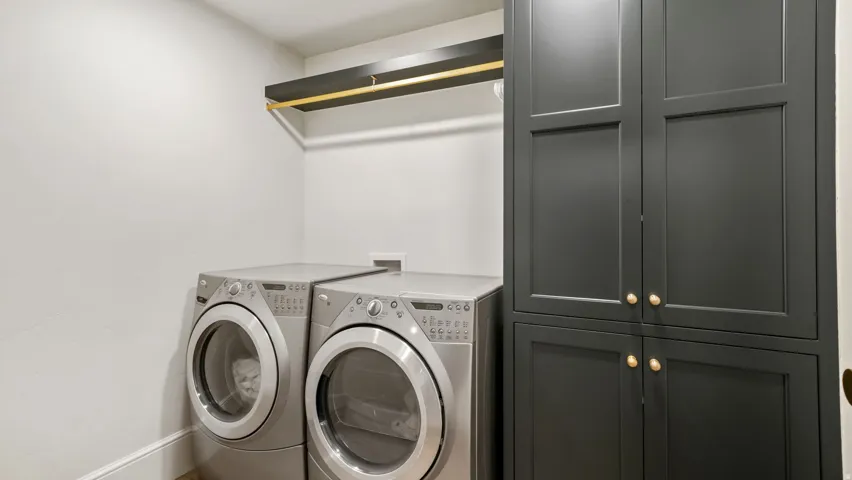 Laundry area featuring built-in dark cabinetry with gold-toned hardware, a wall-mounted shelving unit with a gold-toned hanging rod, and light-toned walls