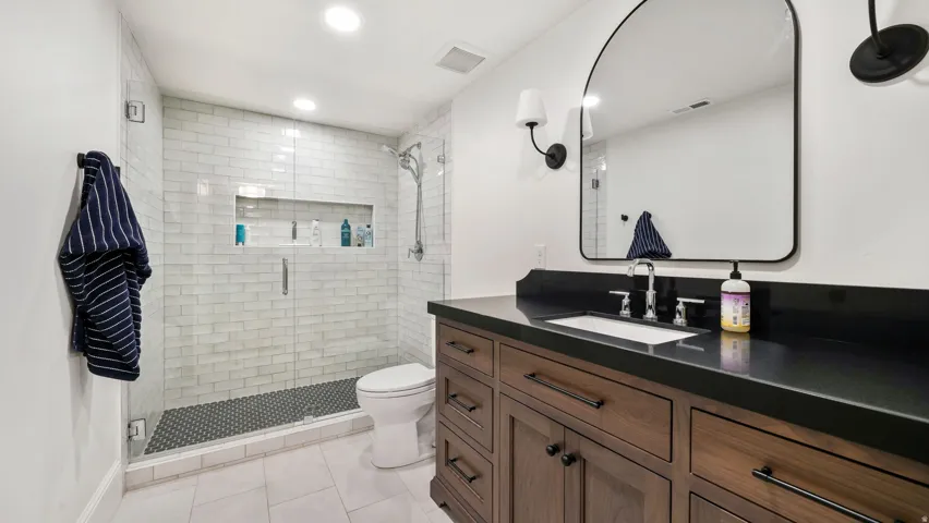 Bathroom featuring a dark wood vanity with a dark countertop, an arched mirror, and a glass-enclosed shower with white subway tile and a black hexagonal tile floor