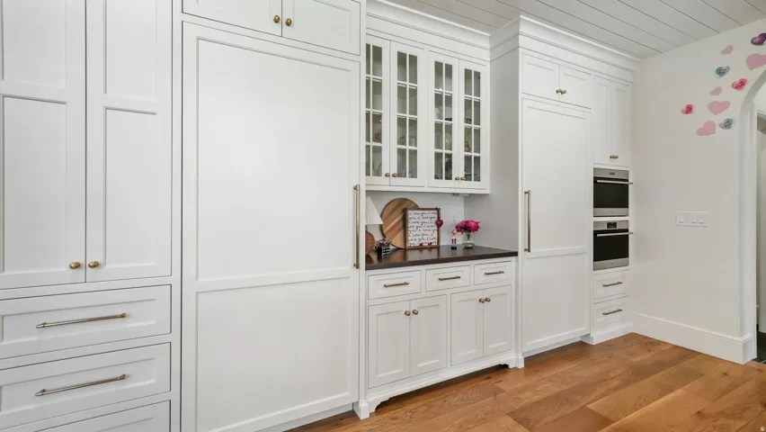 Built-in refrigeration and double ovens framed by custom white cabinetry with glass-front uppers, brass hardware, and a dark stone countertop