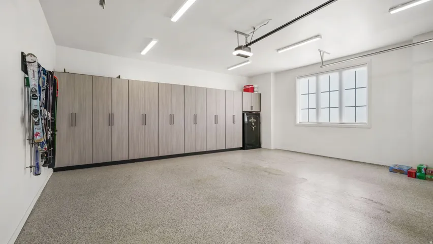 Spacious garage featuring extensive built-in cabinetry, epoxy flooring, ample natural light from a large window, and overhead strip lighting
