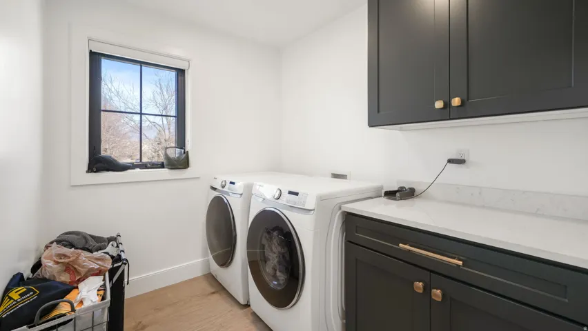 Dedicated laundry room featuring white walls, wood-finish flooring, a black grid-pattern window, dark cabinetry with gold hardware, and a light-colored countertop