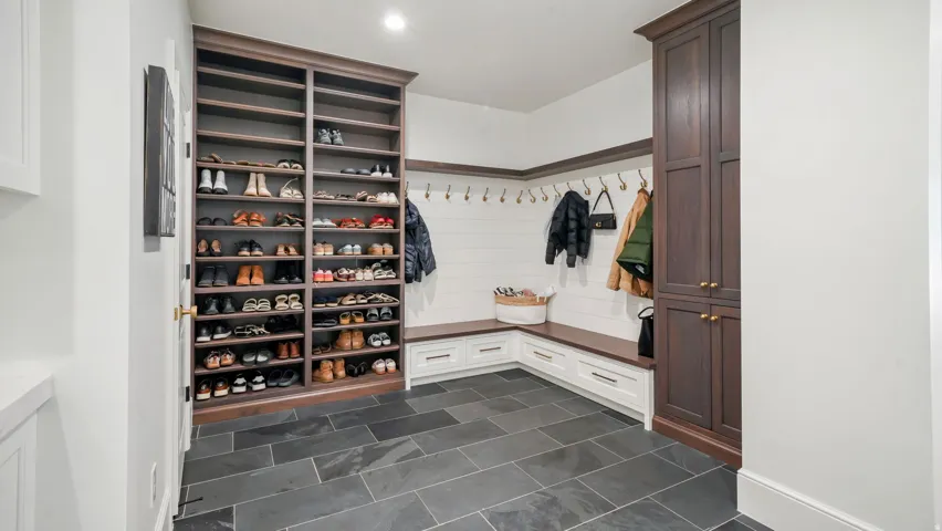 Spacious mudroom featuring dark rectangular floor tiles, built-in shoe shelving, custom wood cabinetry, a shiplap accent wall, and a corner bench with integrated storage drawers