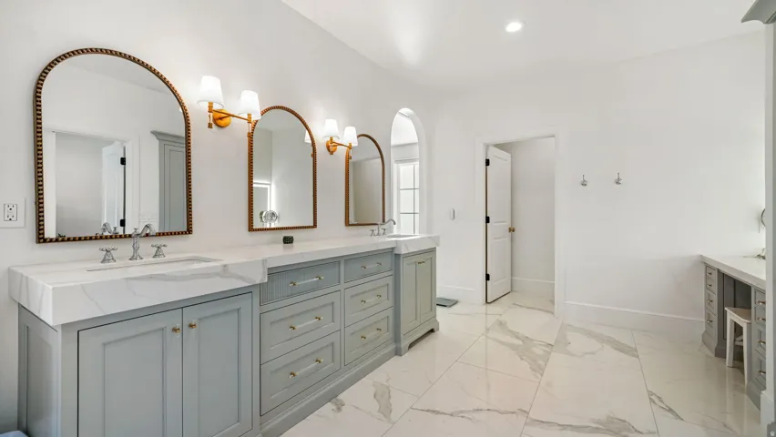 Spacious bathroom featuring a dual vanity with light gray cabinetry and white countertops, arched mirrors, brass wall sconces, and large format white tile flooring with gray veining