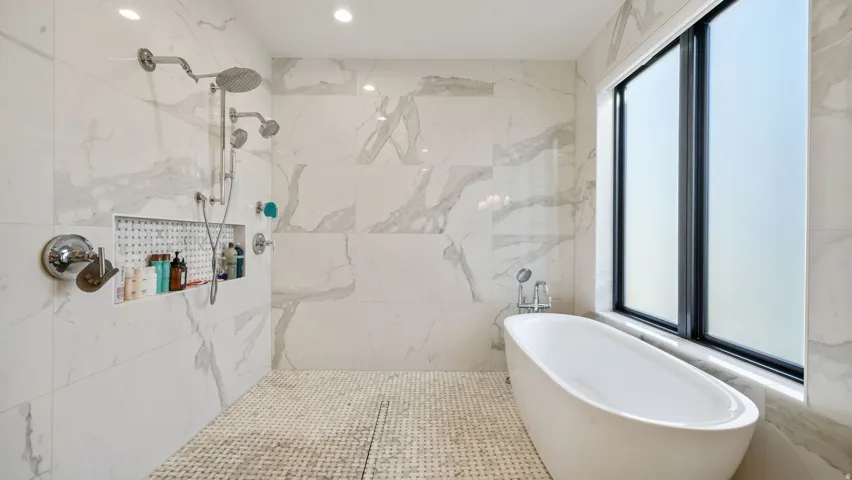Freestanding soaking tub with a large privacy window, complemented by a spacious walk-in shower featuring multiple showerheads, a built-in niche, and marble-look wall tiling