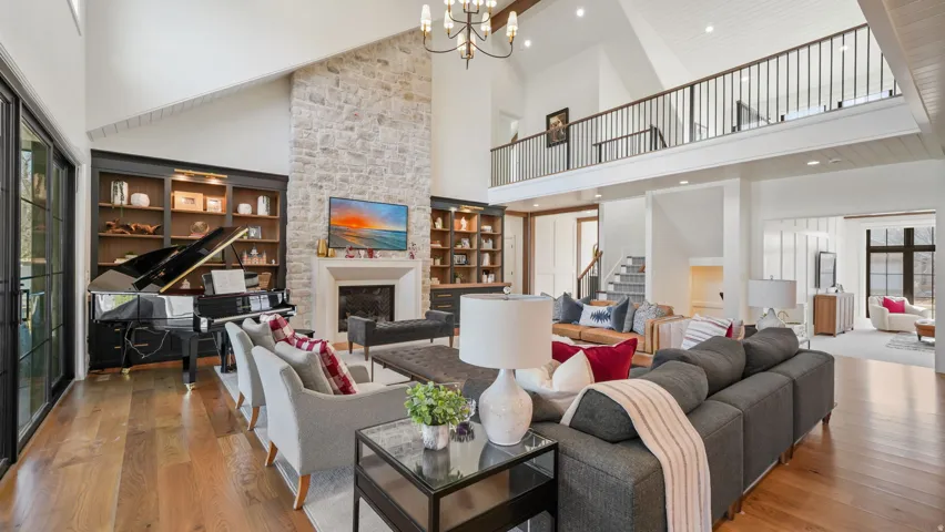 Spacious living area featuring a two-story stone fireplace, vaulted ceilings with wood beams, wood-finish flooring, built-in cabinetry, and a second-story interior balcony with metal railings