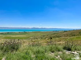 Expansive landscape featuring a large body of water, rolling green hills, a clear blue sky, and distant mountains