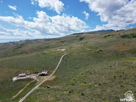 Expansive rolling hills with natural vegetation, featuring a distant body of water, a winding dirt road, and a remote building site
