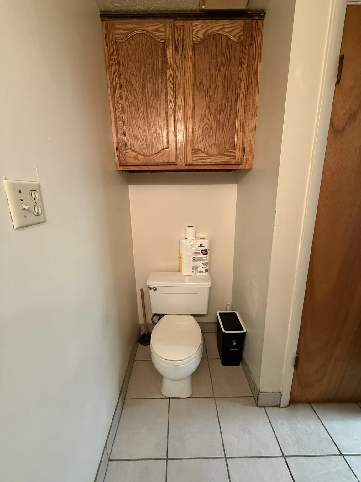 Recessed toilet area featuring a white toilet, overhead wood-finish cabinetry, and light-toned square tile flooring