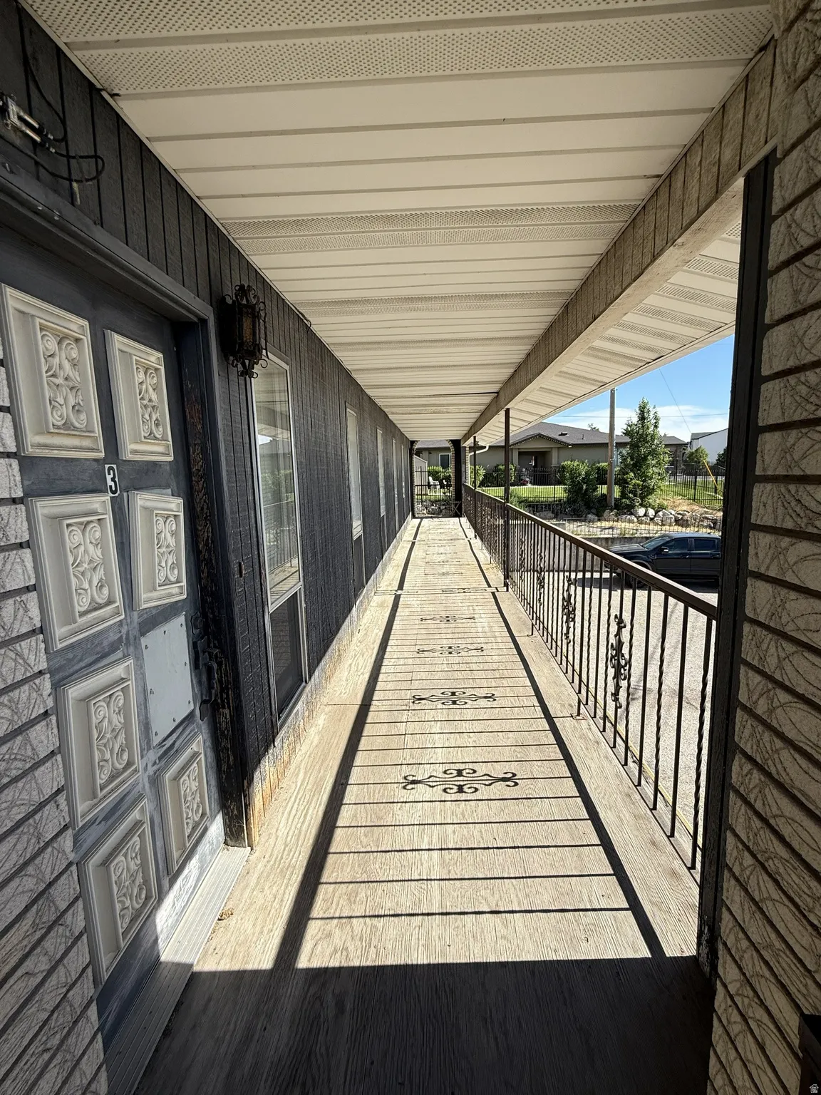 Covered exterior walkway featuring light wood-finish flooring with decorative inlays, dark metal railing, and a black paneled door with ornate white detailing