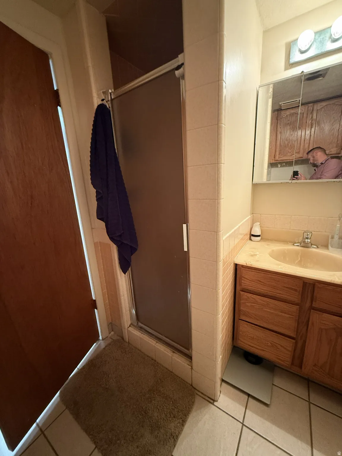 Bathroom featuring a tiled shower enclosure with a frosted glass door, a vanity with wood-finish cabinetry, a single basin sink, a wall-mounted mirror, and tile flooring
