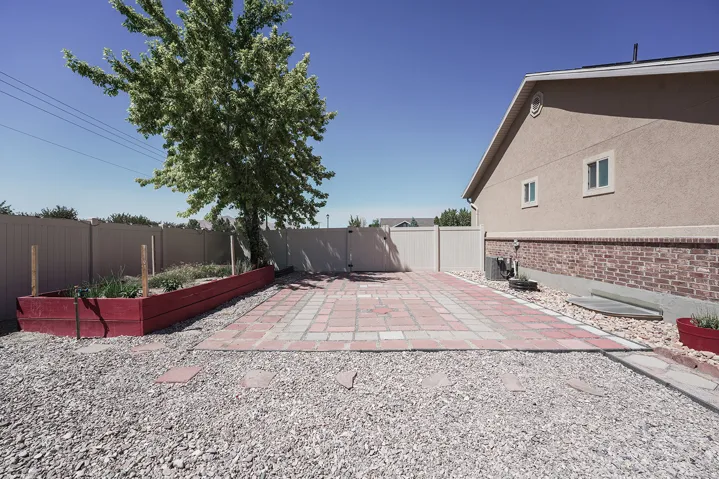 Paved patio area with brick and stucco exterior, featuring a privacy fence, mature tree, and raised garden bed