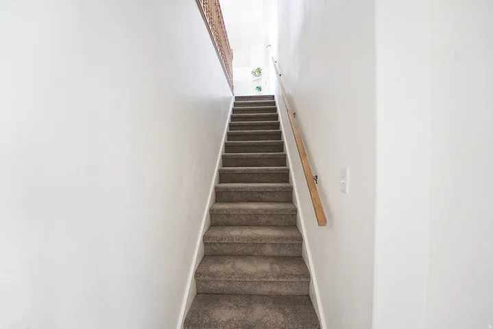 Carpeted staircase with light wood handrail, white wall finishes, and a partial view of a decorative metal balustrade