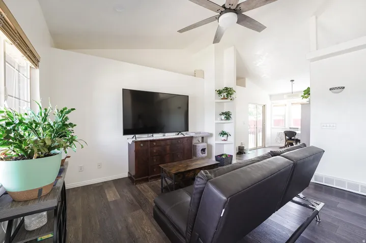 Living area featuring vaulted ceilings, wood-finish flooring, a ceiling fan, built-in corner shelving, and large windows