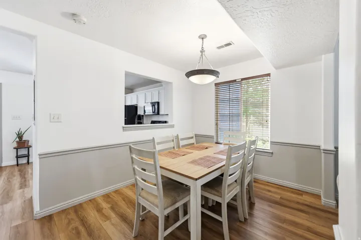 Dining area featuring wood-finish flooring, a contemporary ceiling light fixture, a window with blinds, and a pass-through opening to the kitchen