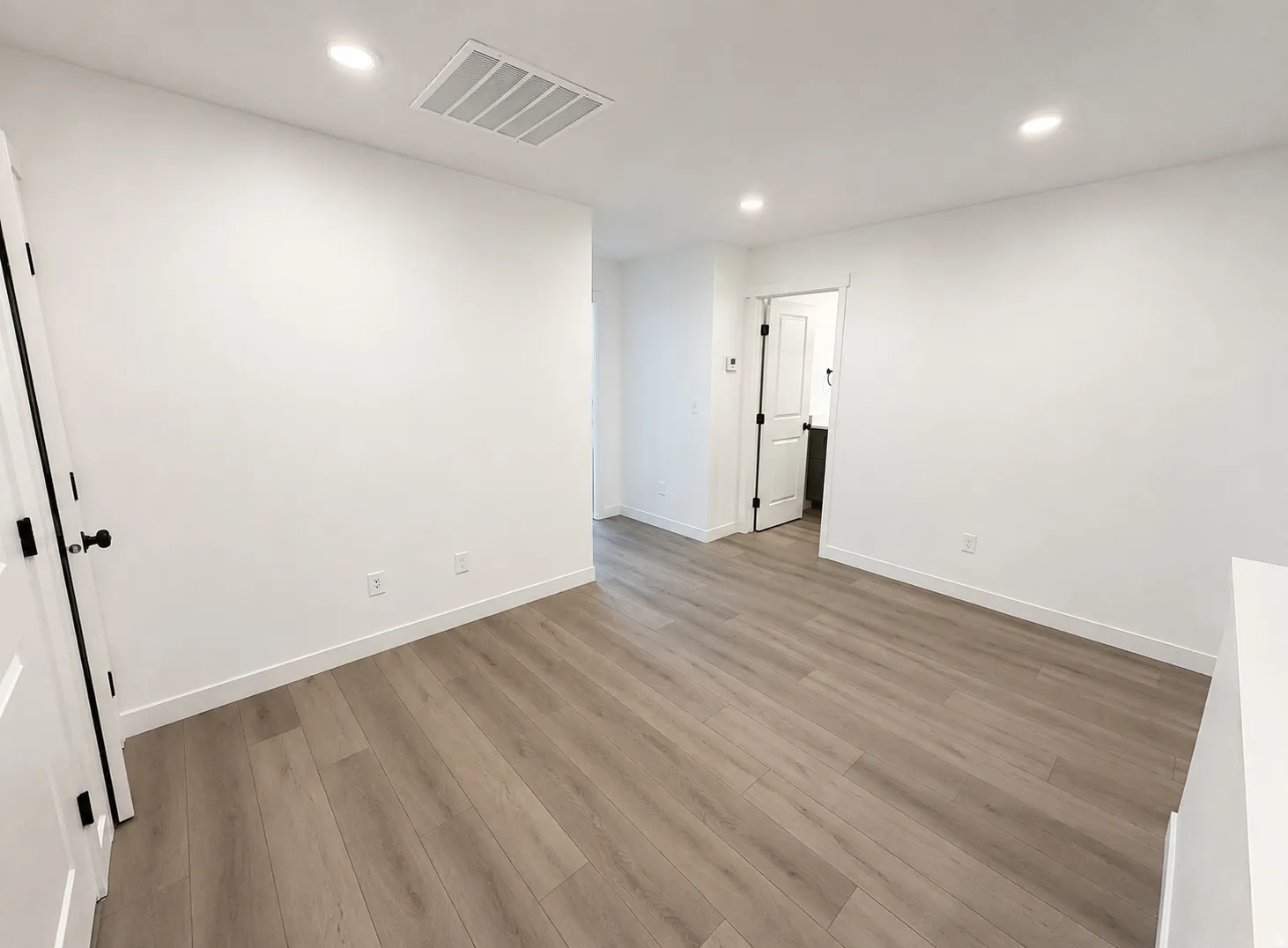 Clean white walls, recessed lighting, wood-finish flooring, white door with black hardware, and an open doorway