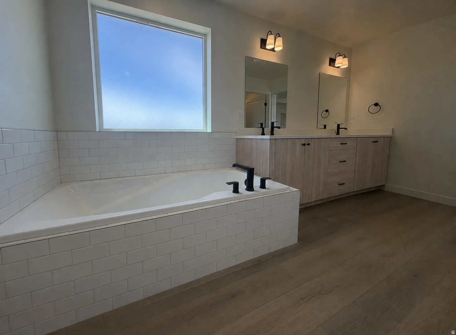 Soaking tub with white subway tile surround, dual vanity with wood-finish cabinetry, light countertops, matte black fixtures, and wood-finish flooring