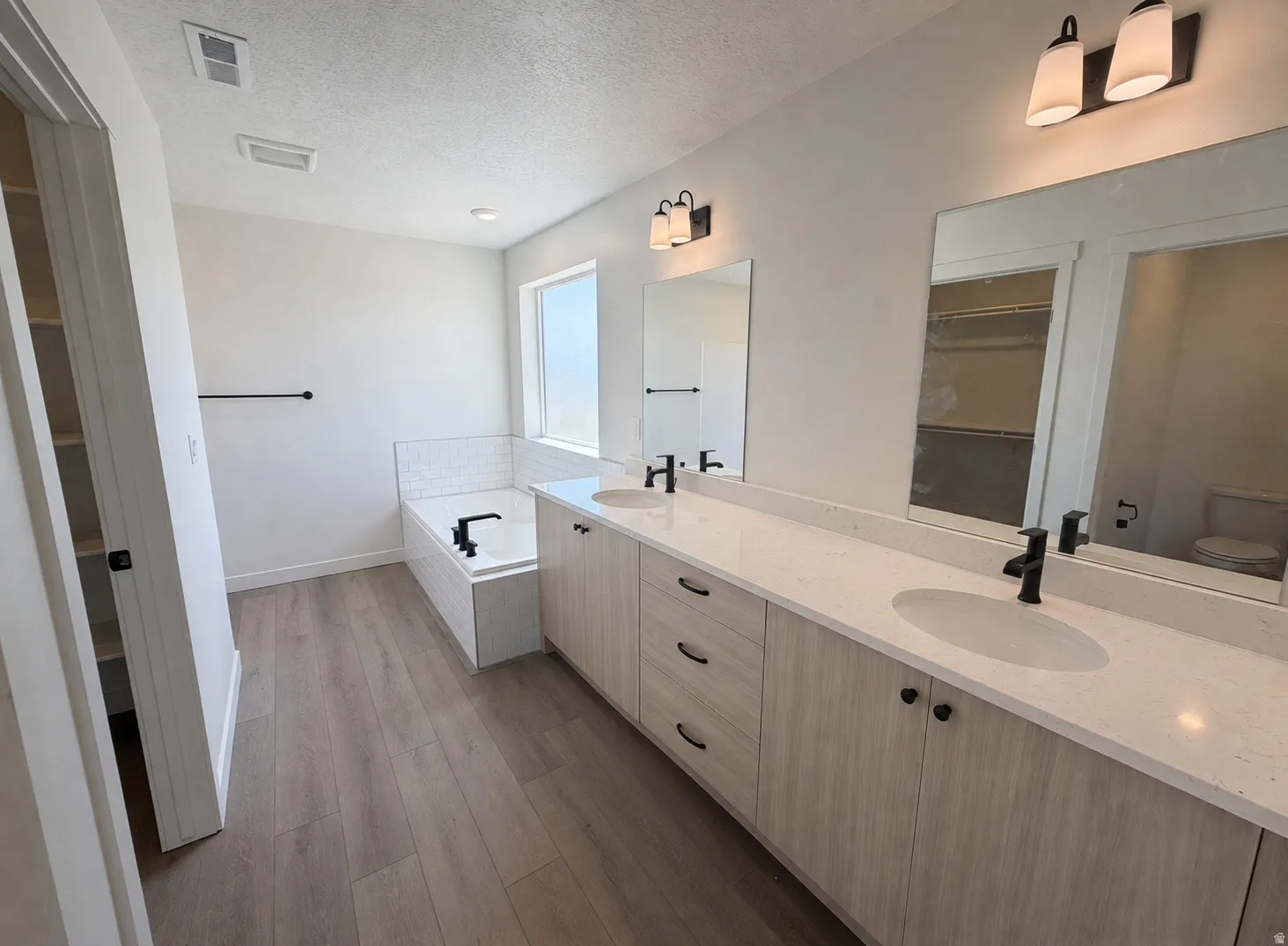 Dual vanity with integrated sinks and black fixtures, featuring a built-in soaking tub with subway tile surround, wood-finish flooring, and a large window across from a shower