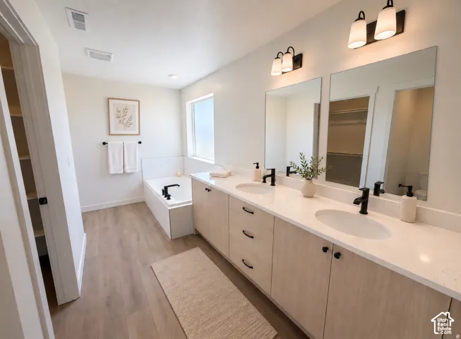 Dual vanity with integrated sinks, light wood-finish cabinetry, black hardware, built-in bathtub with tiled surround, and wood-finish flooring