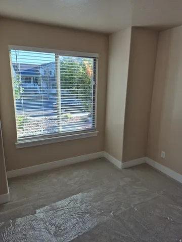 Carpeted interior space featuring a large window with blinds, beige painted walls, and white baseboards
