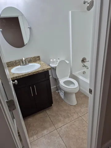 Bathroom featuring a dark wood-finish vanity with a light speckled countertop and undermount sink, an oval wall-mounted mirror, a white toilet, and a combined tub and shower with a rainfall showerhead