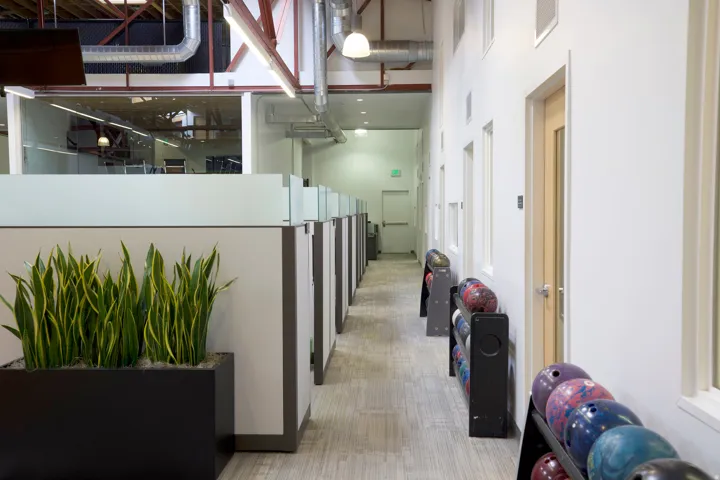 Hallway with cubicle workstations featuring frosted glass partitions, exposed industrial ceiling, and neutral-toned carpet flooring