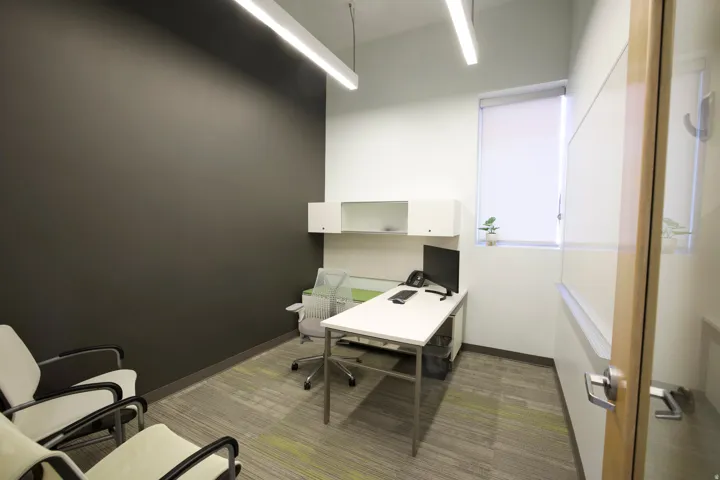 Office space featuring modular carpet flooring, recessed built-in shelving, a window with a roller shade, and linear fluorescent lighting fixtures