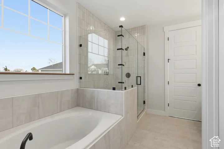 Tiled soaking tub featuring a large window, glass-enclosed shower with matte black fixtures, light-toned floor tiling, and a modern five-panel interior door